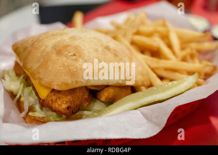 Deep fried fish sandwich on sour dough bun with cheese and lettuce.  Sandwich and fries in red basket with white paper liner.  Pickle included. Stock Photo