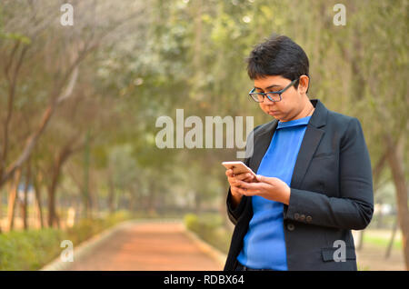 Closeup portrait of confident young Indian Corporate professional woman with short hair, checking her phone in an outdoor setting wearing a black suit Stock Photo