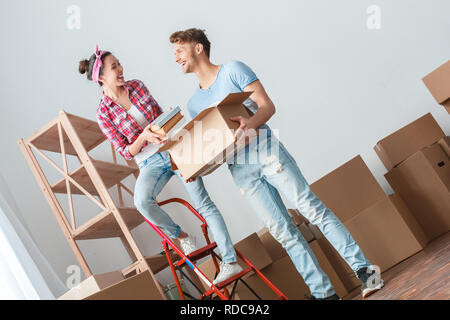 Young couple moving to new place woman standing on ladder putting books into box laughing cheerful with man Stock Photo