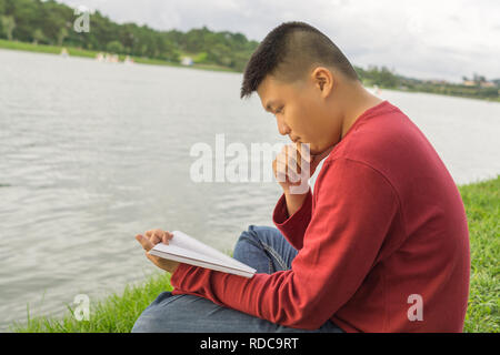 Asian man sitting on grass, reading book Stock Photo