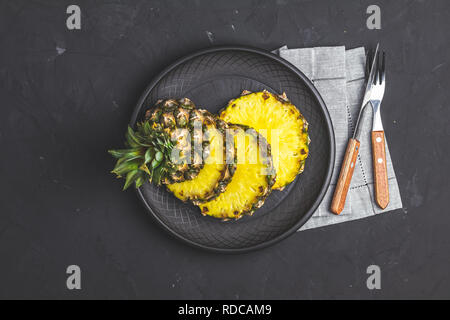 Sliced pineapple in ceramic plate and set of cutlery knife, fork  on black stone concrete textured surface background. Top view with copy space for yo Stock Photo