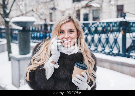 Charming smiling young woman in fur coat with coffee walking in snowfall in Europe city centre. Expressing positivity, true emotions, enjoy snowing, w Stock Photo