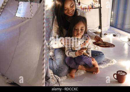 Photo of amazing asian family mother and daughter eating cookies while resting together at home in children playing tent Stock Photo