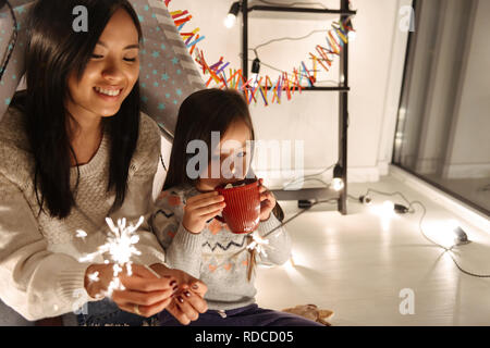 Photo of a happy young asian woman with her little daughter girl sitting on floor holding bengal lights. Christmas concept. Stock Photo