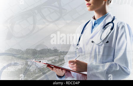Portrait of young female doctor in white sterile suit holding notebook in hands while standing against modern city view and medical equipment on backg Stock Photo