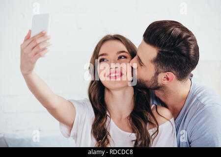 boyfriend kissing girlfriend while taking selfie in bedroom Stock Photo