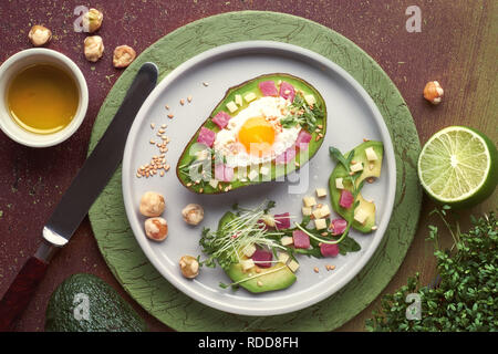 Keto diet dish: baked Avocado boat with ham cubes, quail egg and cheese served with green rucola salad, flat lay with ingredients Stock Photo