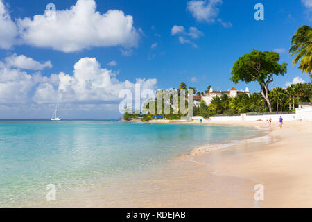 A yacht anchors off Gibbes Bay, an empty beach with palm trees and an exclusive resort at its point. A few people meander along the beach. Stock Photo