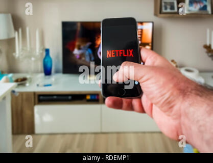 Rome, Italy, 12 January 2019; Human hand uses the smart phone to watch Netflix's streaming content with a TV set in the background Stock Photo