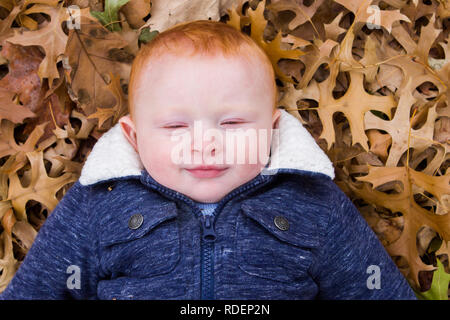 Close up Red headed little boy lying down in leaves outside Stock Photo