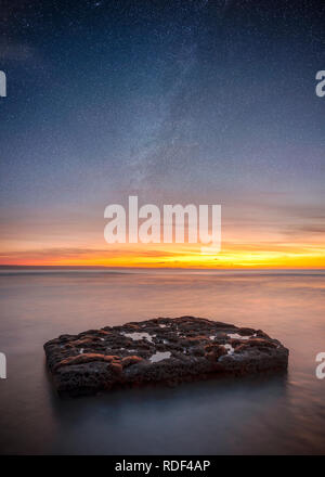 long exposure of a rock in the sea with sun setting in distance revealing the milky way Stock Photo