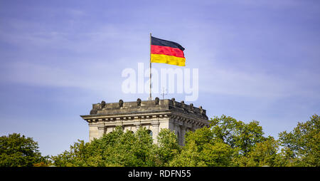 German flag waving on silver flagpole, Reichstag building in Berlin. Blue sky with clouds background, trees in front of building. Stock Photo
