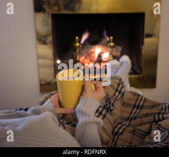 Close up image of woman sitting under the blanket by cozy fireplace warming up her feet in woolen socks relaxing with cup of hot chocolate drink at ho
