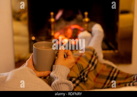 Close up image of woman sitting under the blanket by cozy fireplace warming up her feet in woolen socks relaxing with cup of hot chocolate drink at ho