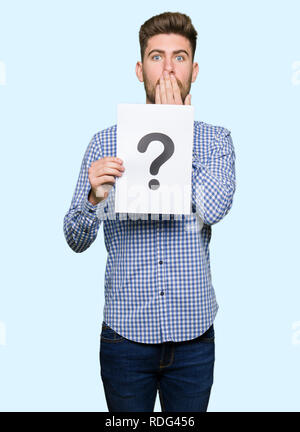 Young handsome man holding paper with question mark cover mouth with hand shocked with shame for mistake, expression of fear, scared in silence, secre Stock Photo