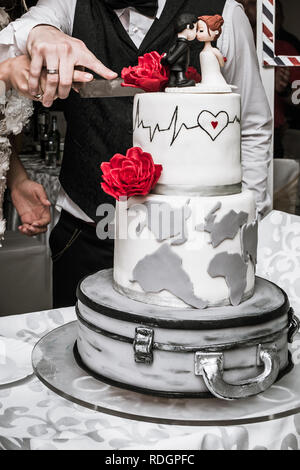 Bride and groom cut wedding cake with red roses, heartbeat rate and figurines on the table in restaurant. Stock Photo