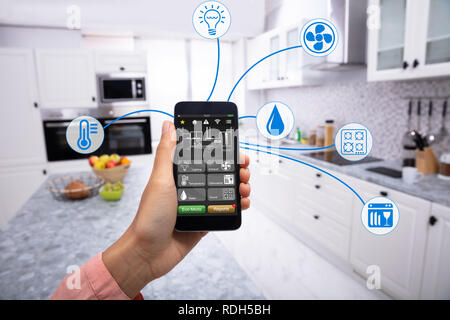Woman's Hand Using Home Control System On Cellphone With Various Icons In The Kitchen Stock Photo