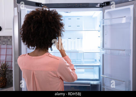 Rear View Of Worried Woman Looking At Empty Refrigerator In Kitchen Stock Photo
