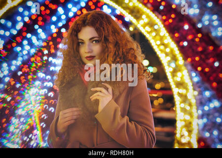 Young attractive red-haired girl. Portrait on the night street of the city. Bokeh from festive lights. Stock Photo