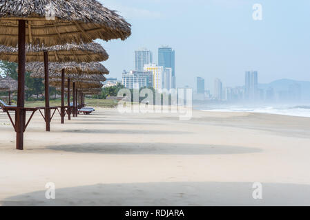 Danang, Vietnam - November 1, 2018: Thatched umbrellas along the beach with the Da Nang city skyline. Stock Photo