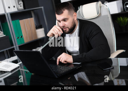 Busy young man talking on phone and looking at laptop in office Stock Photo