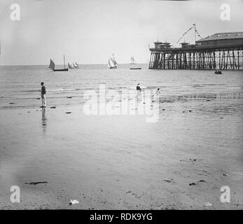 Late Victorian photograph showing the beach and the end of the Pier in Brighton, England. Some small sail boats on the sea, and a few people enjoying the beach. Stock Photo