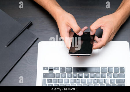 Mans hands typing on smart phone while working on laptop computer on vintage wooden table. Hands typing on a keyboard. Top view, business office workp Stock Photo