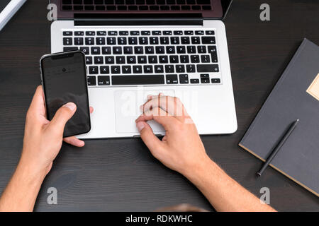 Mans hands typing on smart phone while working on laptop computer on vintage wooden table. Hands typing on a keyboard. Top view, business office workp Stock Photo