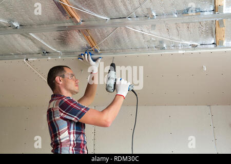 Young man in goggles fixing drywall suspended ceiling to metal frame using electrical screwdriver on ceiling insulated with shiny aluminum foil. Renov Stock Photo