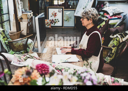 Senior woman wearing glasses, red dress and white apron sitting at wooden table in a studio, working on desktop computer. Stock Photo