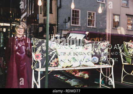 Smiling senior woman wearing glasses and red dress standing in interior design store, looking at camera through window. Stock Photo