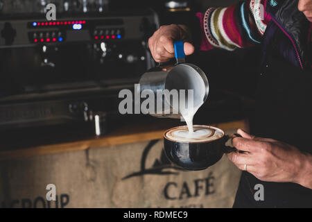Close up of barista pouring milk from jug into cappuccino mug, creating foam pattern. Stock Photo