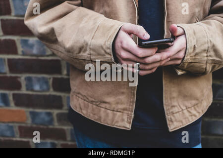 Close up of young man wearing casual jacket standing against a brick wall, texting on his mobile phone. Stock Photo