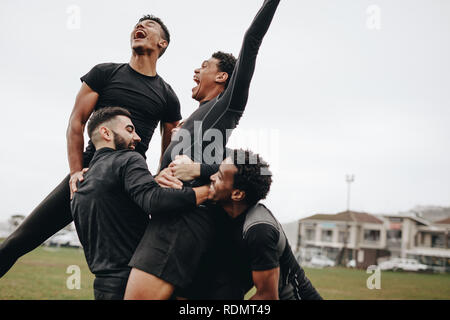 Excited soccer players shouting in joy after scoring a goal. Teammates celebrating victory holding the goal scorer. Stock Photo