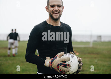 Portrait of a cheerful goalkeeper standing with a soccer ball in hand. Soccer player standing on field holding a football practicing in the morning. Stock Photo