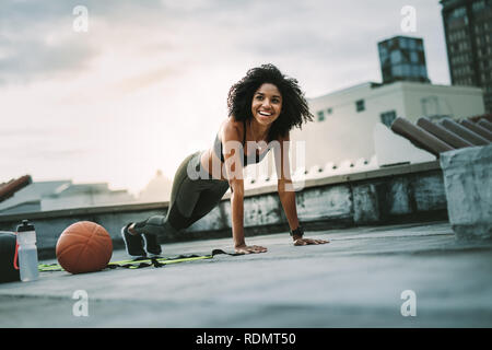 Smiling woman athlete doing push ups on the rooftop with gym balls by her side. Cheerful woman in fitness wear doing workout on the terrace. Stock Photo