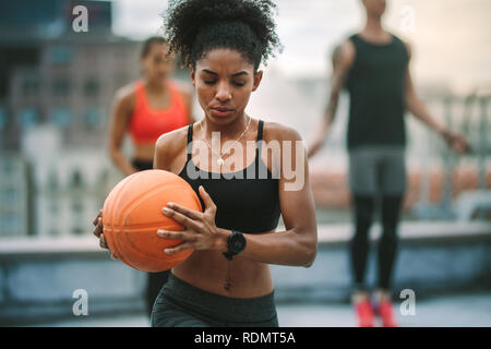Close up of an afro american fitness woman doing fitness training holding a basketball. Female athlete doing workout on rooftop with a man and woman t Stock Photo