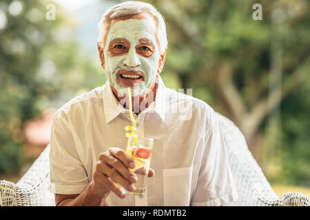 Funny elderly man with clay mask on face drinking juice. Retired senior man at home taking care of his skin at old age. Stock Photo