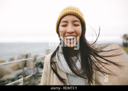 Cheerful asian woman standing beside the sea in warm winter clothes. Happy woman standing outdoors making faces with her hair flying. Stock Photo