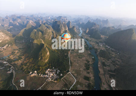 Karst mountains, towns and fields  form backdrop for colorful hot air balloon flying below nearby. Stock Photo