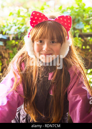 Little readhead girl wearing snowsuit and cat earmuffs and laughing on green leaves background Stock Photo