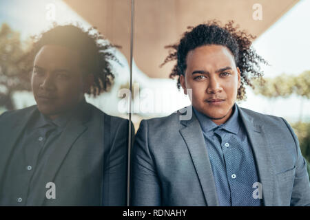 Young mixed race guy with curly afro looking seriously into camera leaning against glass wall Stock Photo