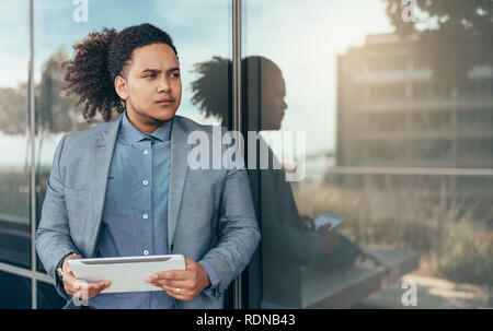 Young mixed race guy with curly afro holding a tablet looking into the distance with reflection on glass wall Stock Photo