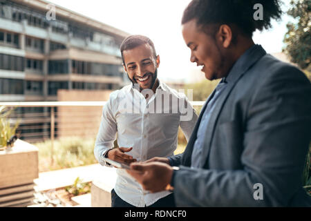 Two businessmen talking and laughing casually outside looking at a digital tablet Stock Photo