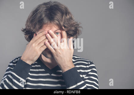 Photo of scared man covers face with both hands, looks through fingers, peeks with fear, has dark curly hair, dressed in comfortable jumper, isolated  Stock Photo