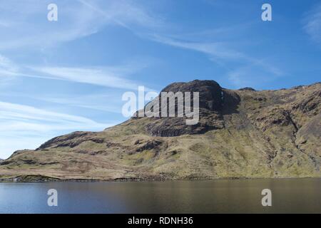 Craggy cliff face of mountain reflected in still water of a mountain lake (tarn). Blue skies and grassy, rocky slopes. Harrison Stickle, overlooking S Stock Photo