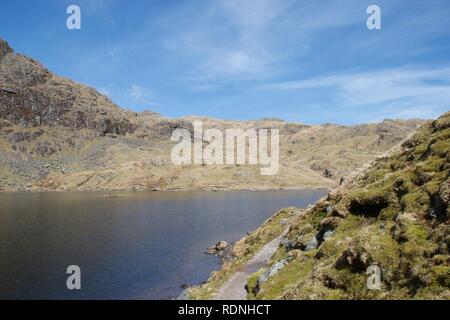 Footpath in foreground, round a clear blue mountain lake (tarn) nestled between craggy, grey, rocky cliffs high on a mountain in the English Lake Dist Stock Photo