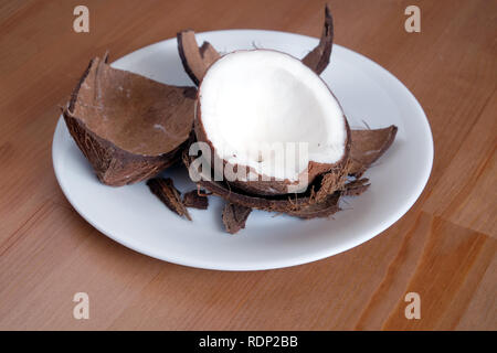 Still life with broken coconut shell ripe white flesh inside and debris on round white plate on brown wooden table horizontal close up photo Stock Photo
