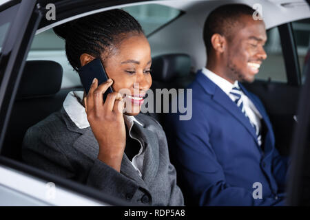 Happy Businesswoman Sitting Inside Car Besides Her Male Colleague Talking On Smartphone Stock Photo