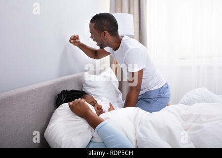 Woman Covering Her Ears With Pillow And Husband Banging Wall For Being Disturbed By Noise Stock Photo